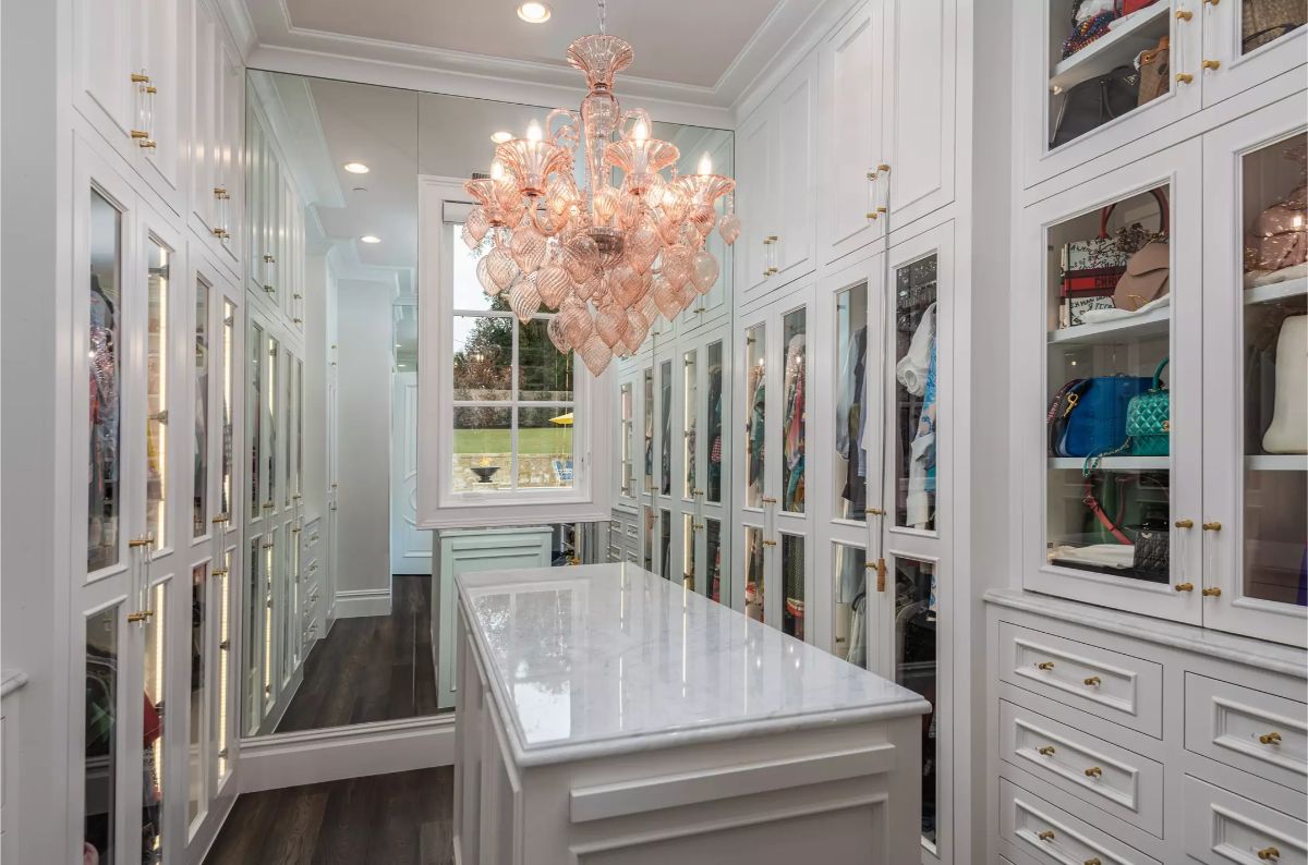 Elegant walk-in closet with white cabinetry, glass-paneled doors, and a central island under a decorative chandelier. 