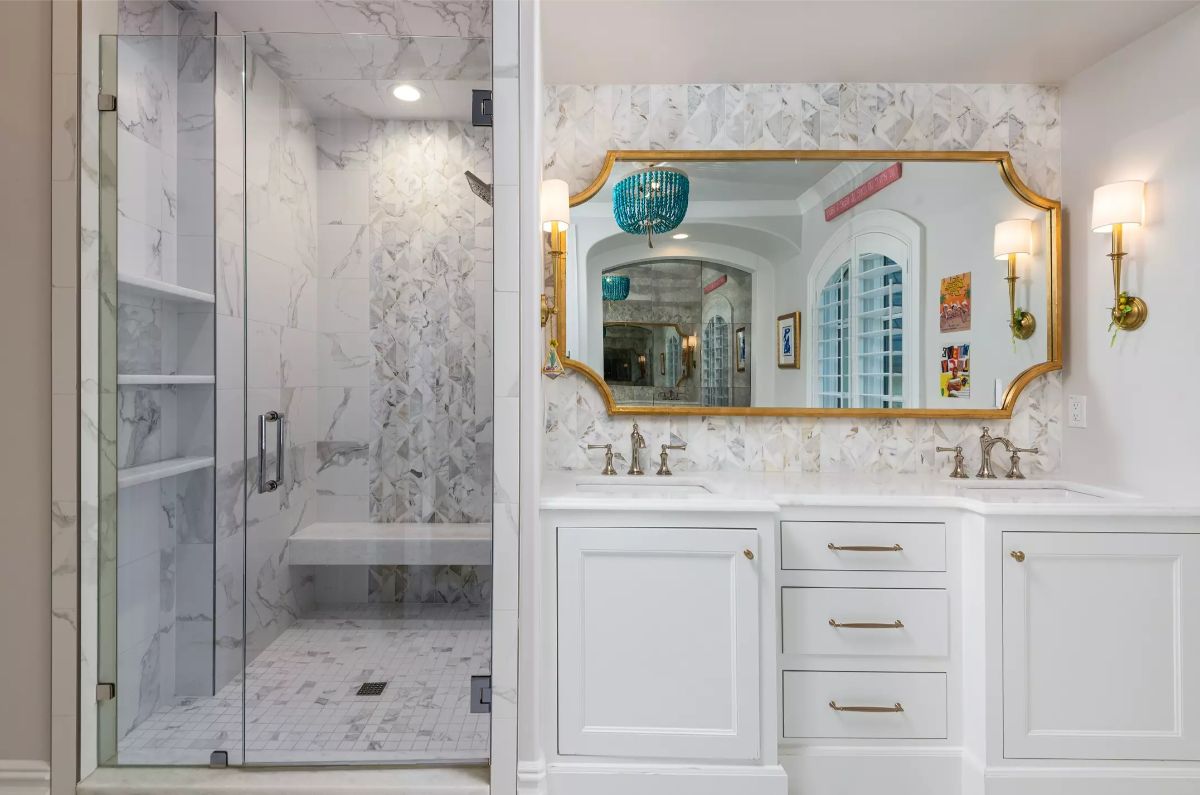 Modern shower and vanity area with marble finishes, a gold-framed mirror, and sleek white cabinetry.