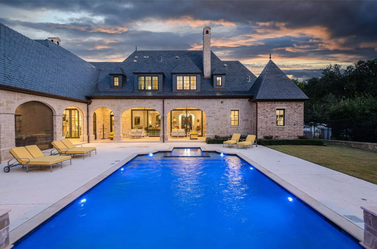 Large pool with glowing blue water surrounded by a stone patio and lounge chairs against a backdrop of a stone house under dramatic evening skies. 