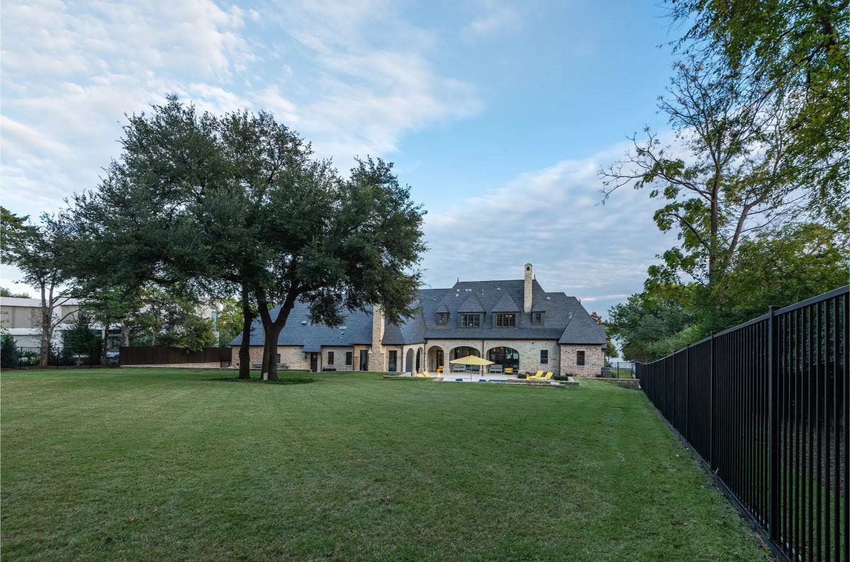 Expansive backyard with manicured lawn, mature trees, and a large stone house featuring a backyard pool and fencing in the distance. 