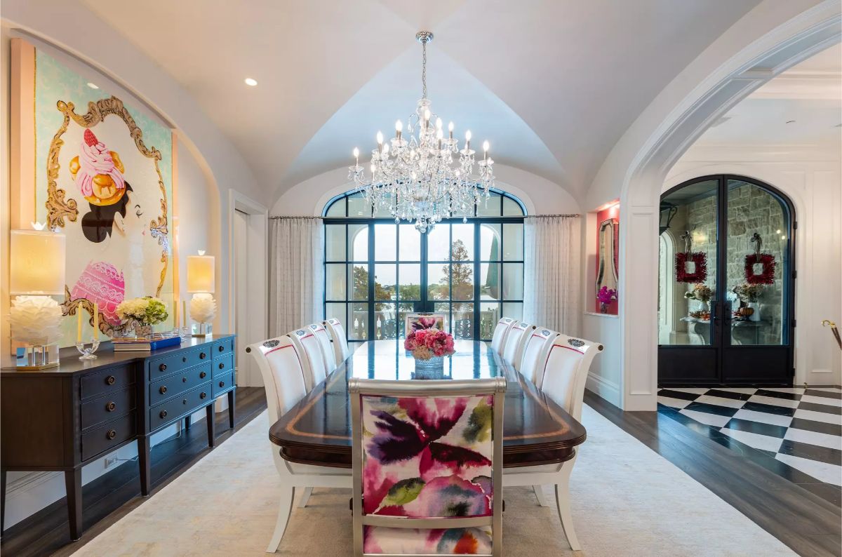 Formal dining room with a long table, crystal chandelier, floral chair accents, and arched glass windows.