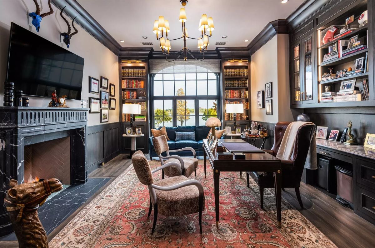 Luxurious library with dark wooden paneling, a fireplace, and a large arched window overlooking a scenic view. 