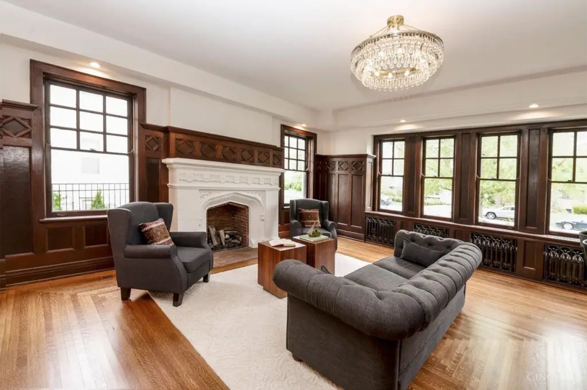 Dark wood paneling lines the walls, adding contrast and depth to the bright space. A white ornate fireplace serves as the focal point of the room, surrounded by comfortable seating.