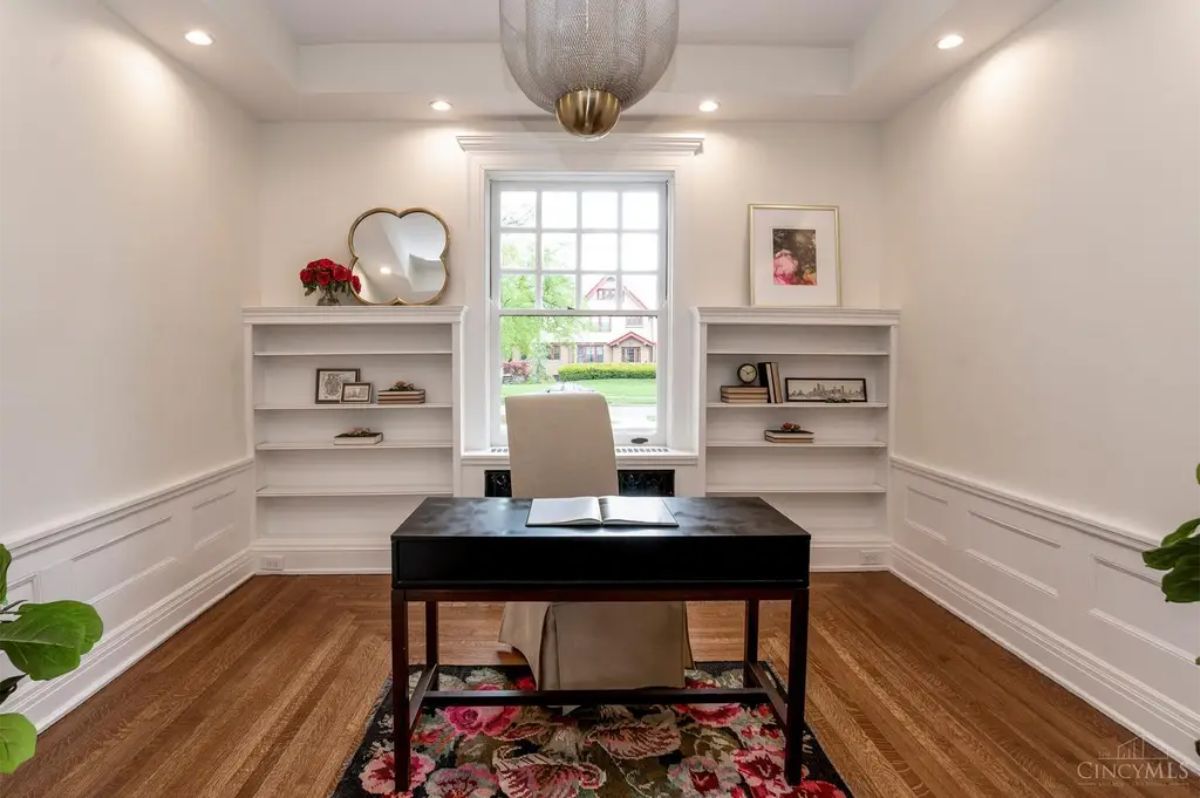 Well-lit home office is centered around a sleek black desk on a floral rug.