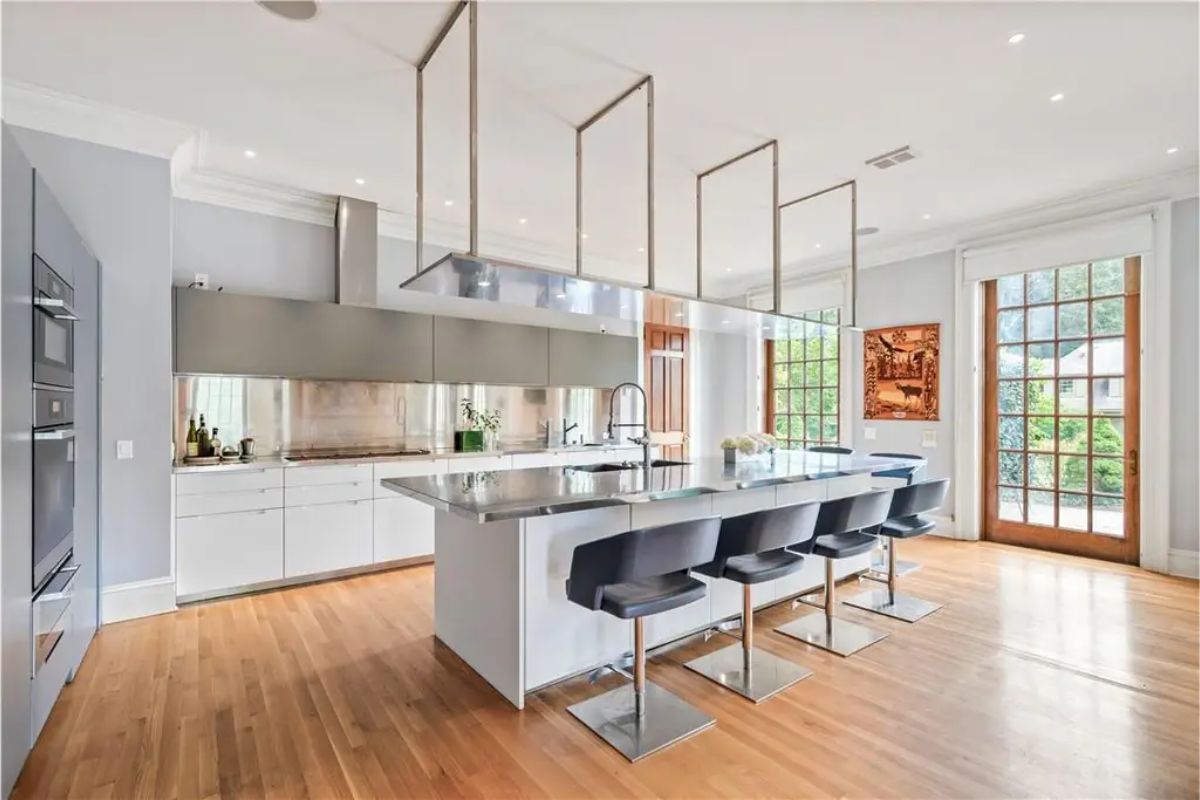 Modern kitchen with a large island, barstools, and a sleek metallic range hood complemented by wooden flooring. 