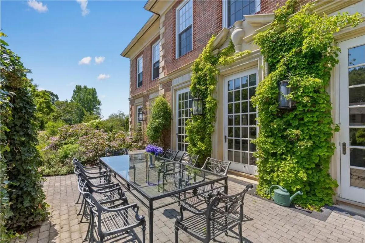 Outdoor patio with a long glass dining table, surrounded by lush greenery and flowering plants. 