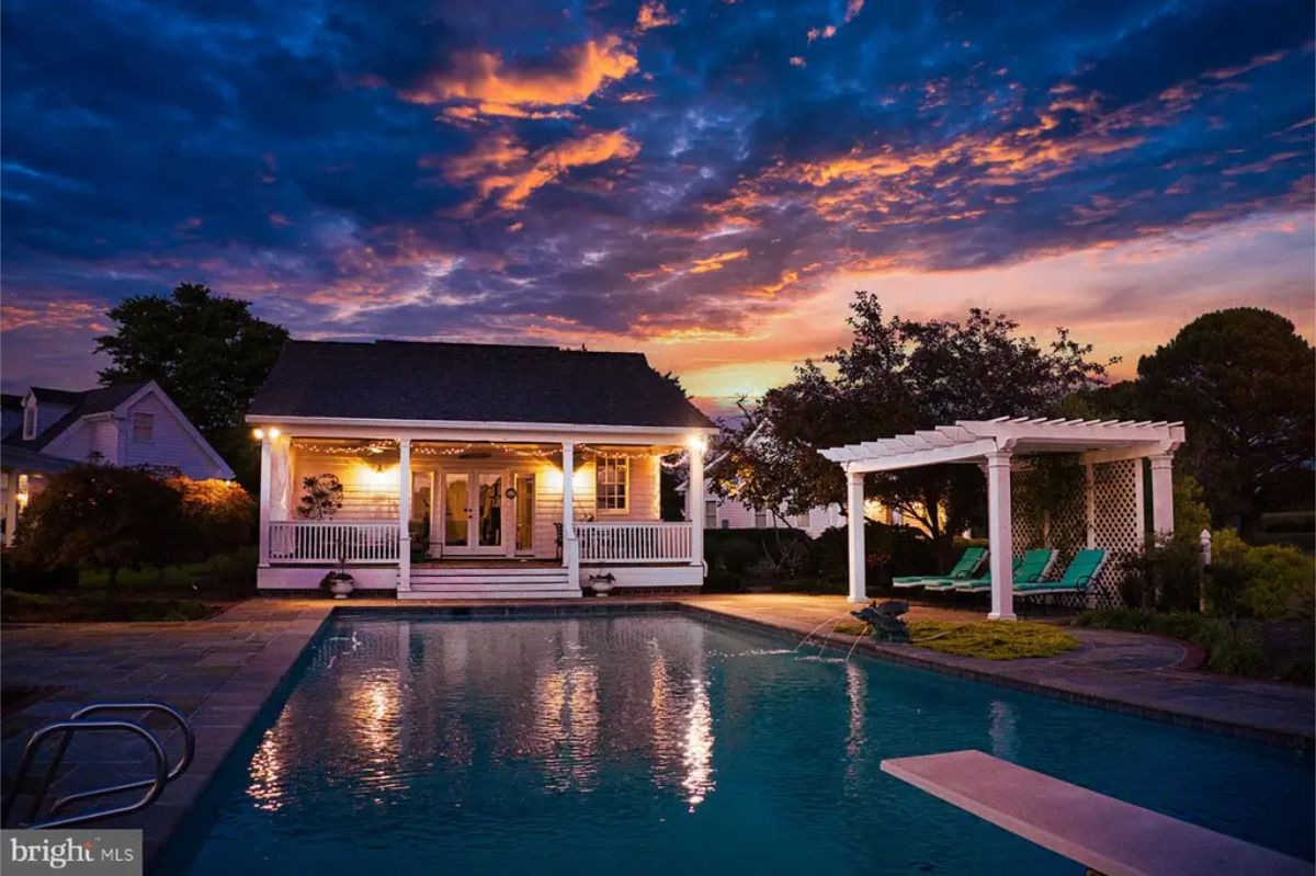 Charming poolside cottage illuminated with warm lighting under a vibrant sunset sky.