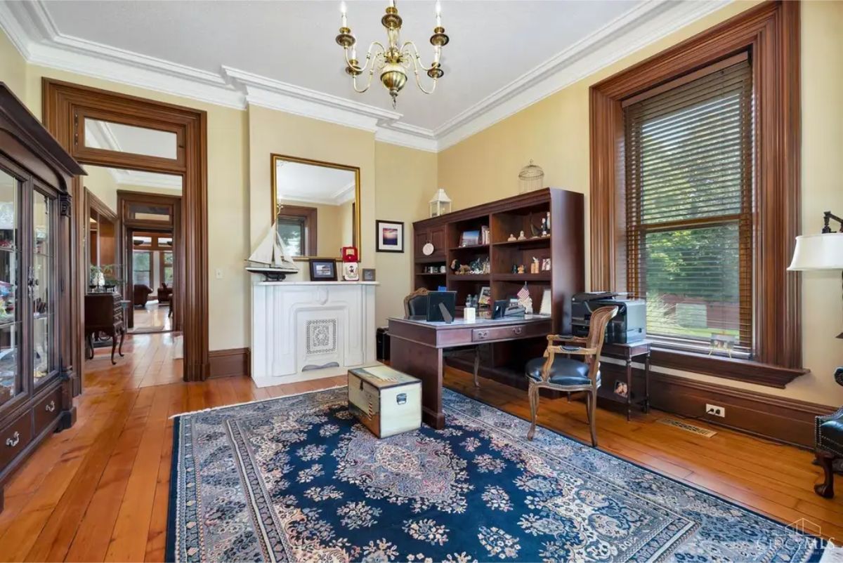Rich wood trim, large windows with natural light, and a decorative white fireplace with intricate detailing.