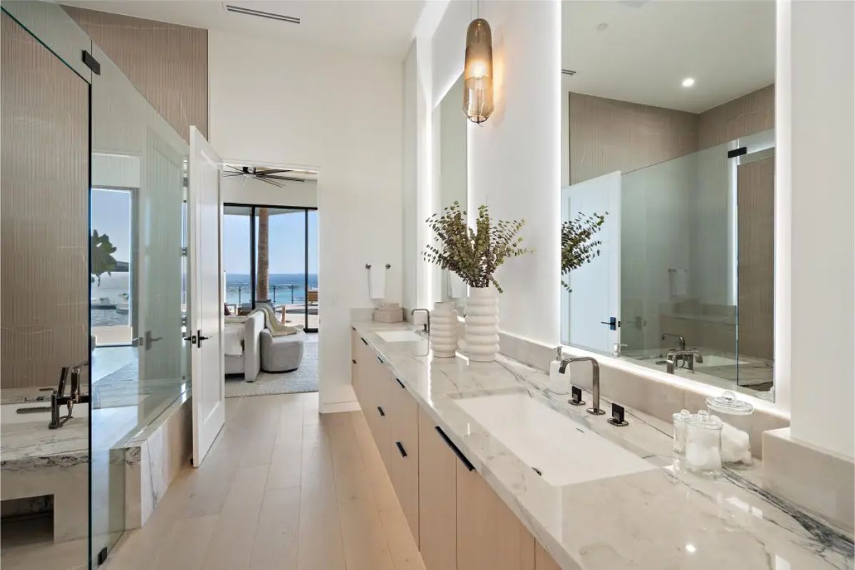 Luxurious bathroom with a double vanity, marble countertops, glass-enclosed shower, and a doorway leading to a bedroom with a poolside view. 