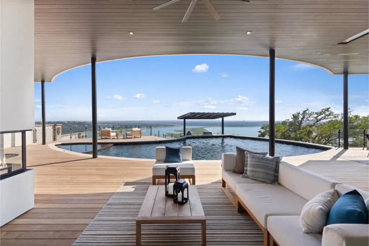 Covered outdoor patio with modern furniture, wooden decking, a ceiling fan, and an infinity pool overlooking a scenic lake. 