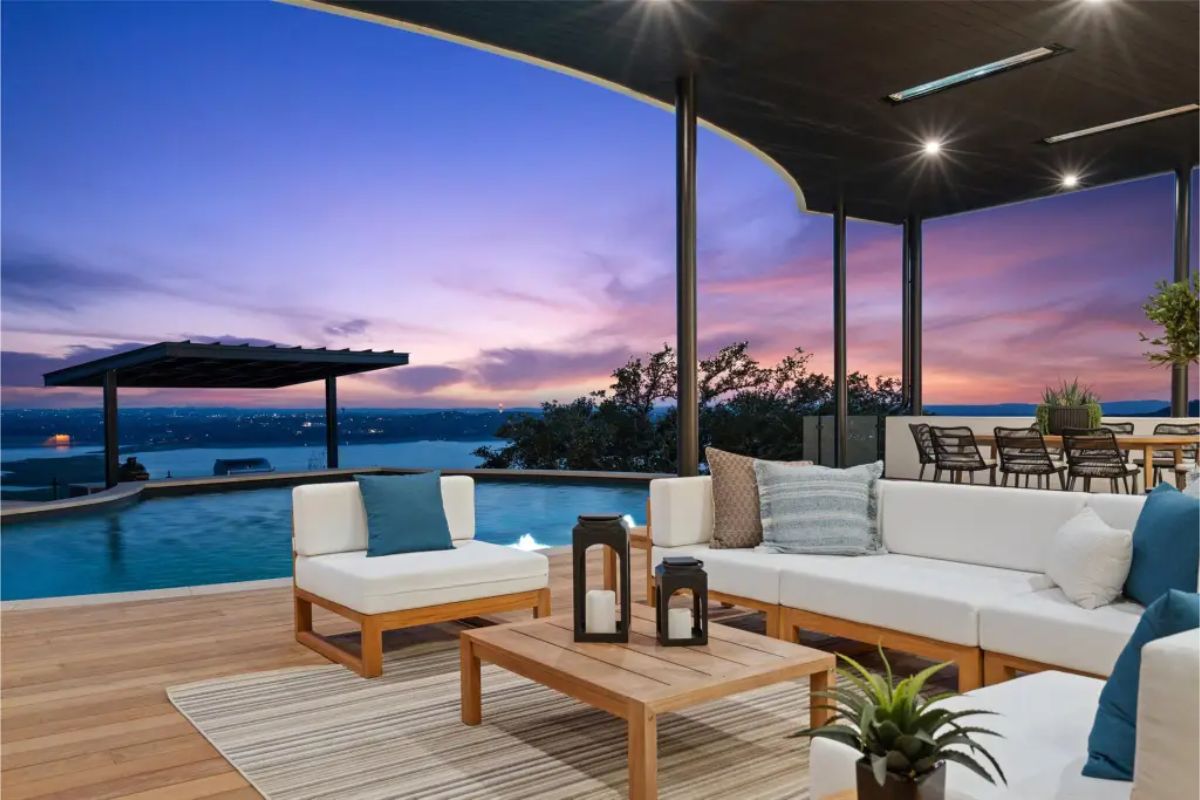 Poolside lounge space at sunset with cozy white seating, a wooden coffee table, and decorative lanterns, set against a backdrop of a vibrant sky. 