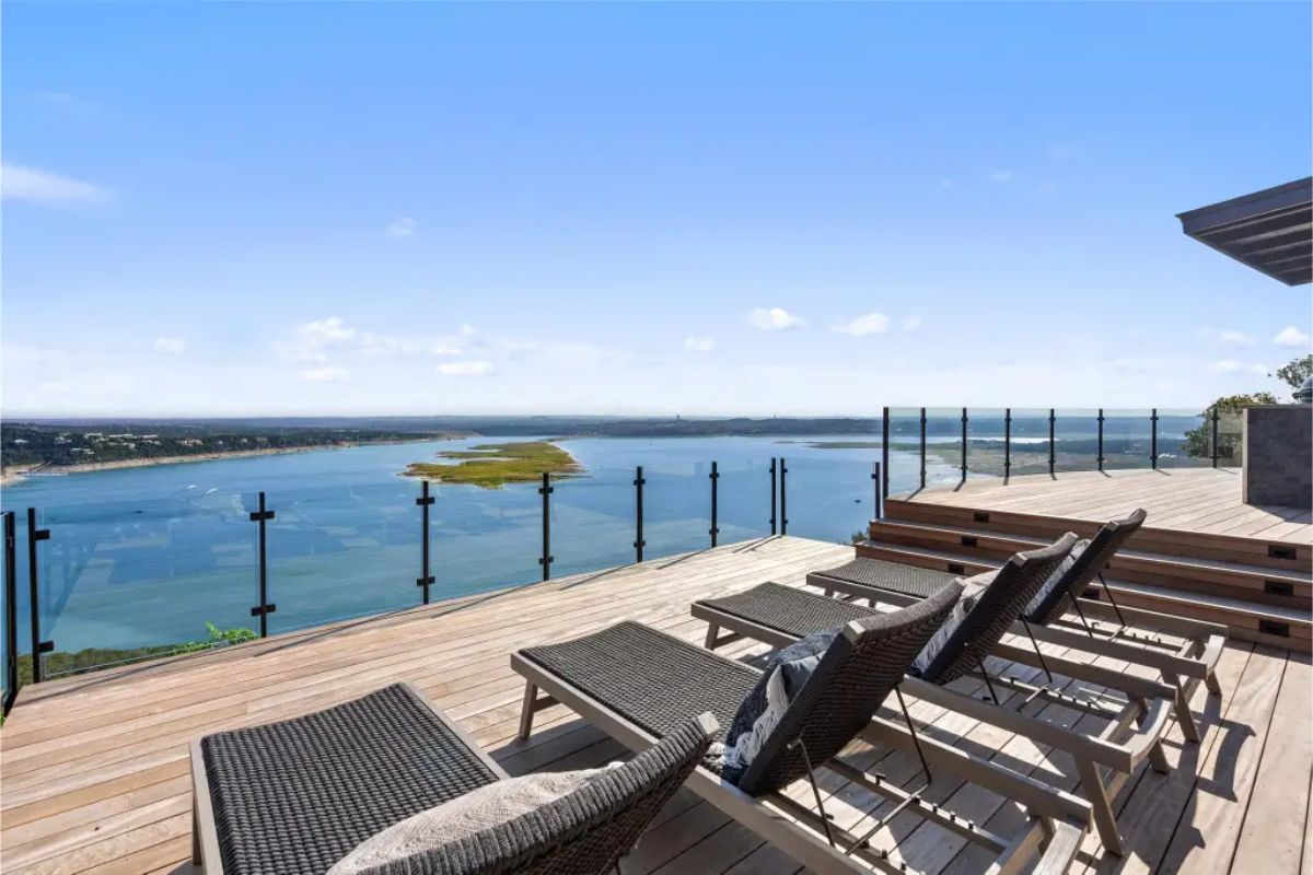 Sunbathing deck with multiple lounge chairs facing a panoramic lake view, enclosed by glass railing. 