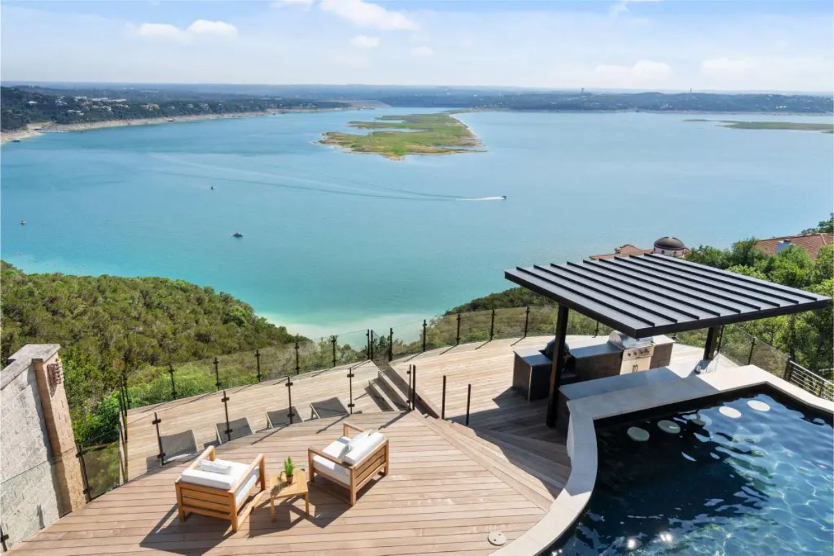 Infinity pool and deck featuring lounge chairs, a glass railing, and an outdoor kitchen with a pergola, all overlooking a picturesque lake. 