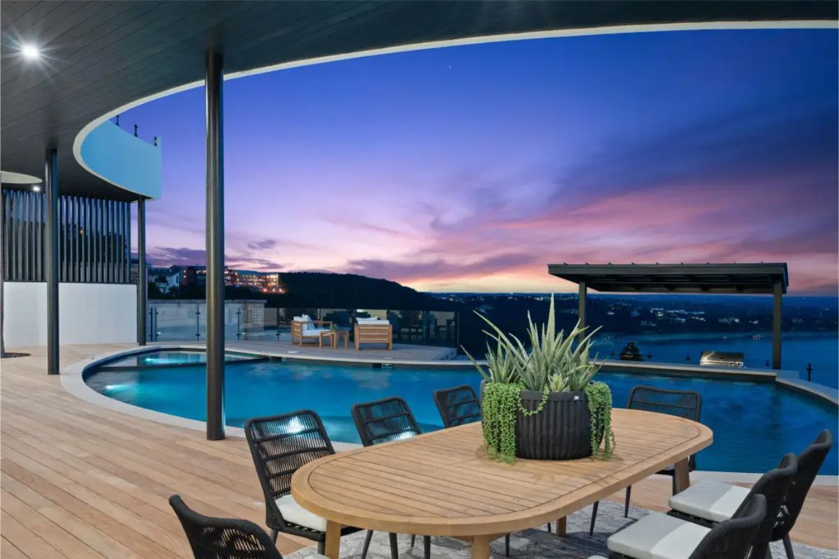Lakeside dining and relaxation area with a large wooden table, modern black chairs, and an infinity pool illuminated by ambient lighting. 
