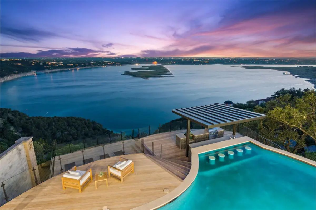 Infinity pool and outdoor entertainment area with a built-in swim-up bar, wooden deck, and breathtaking views of the lake at dusk. 