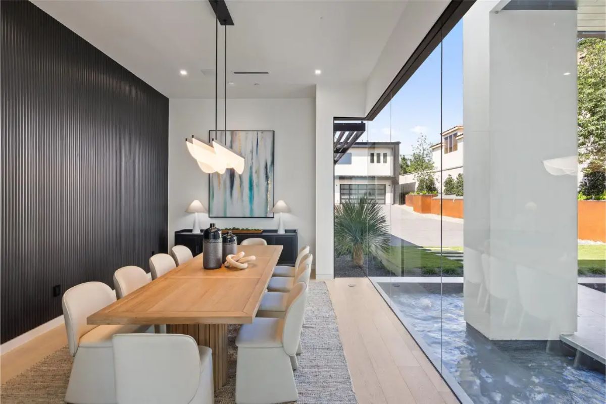 Modern dining room with a wooden table, white chairs, and a floor-to-ceiling glass window overlooking the exterior. 
