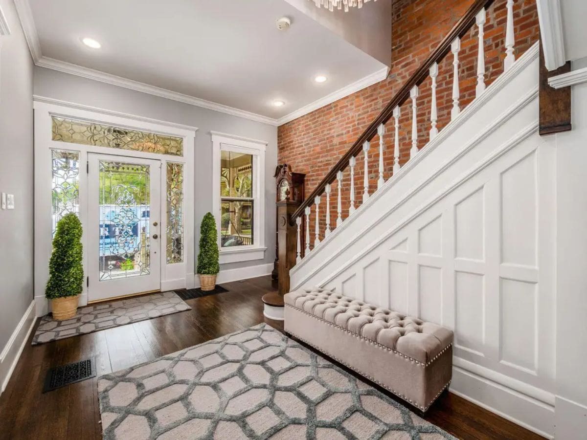 This entryway features a striking blend of historic charm and modern elegance, highlighted by a brick accent wall and a white paneled staircase with detailed balustrades.