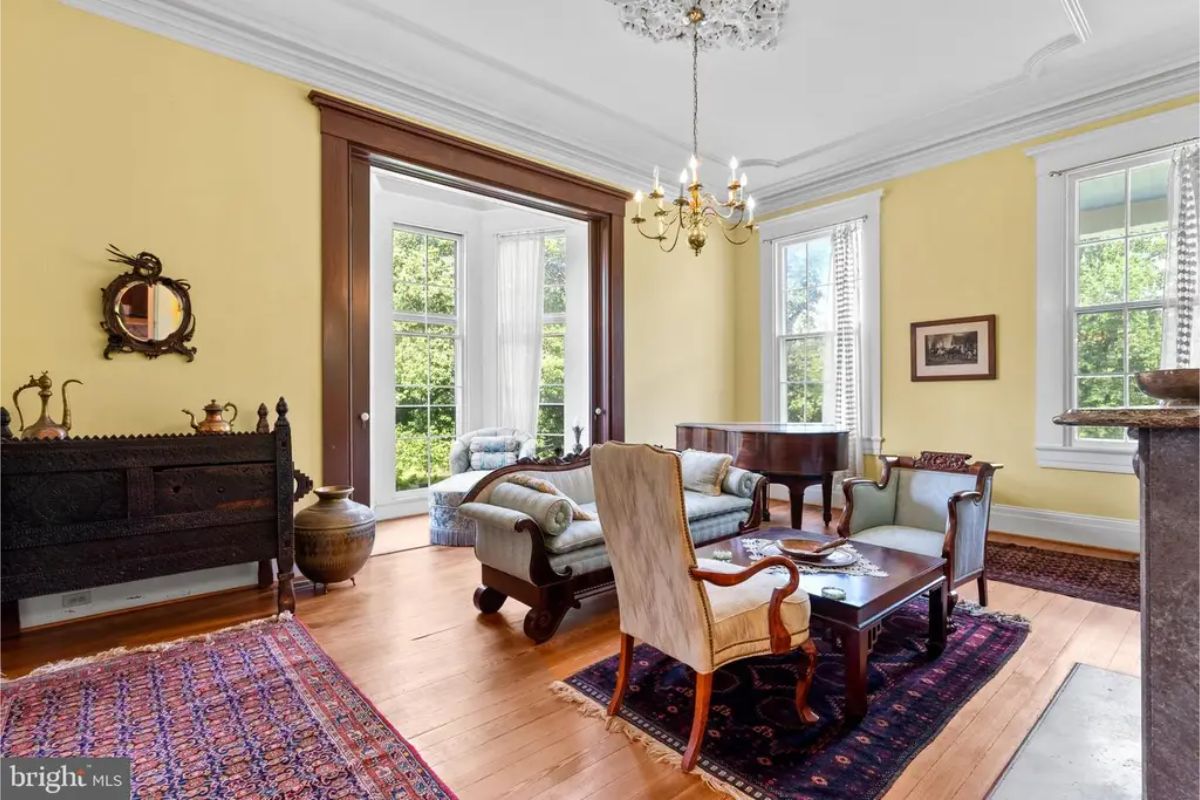Bright room with high ceilings, large windows, and yellow-painted walls. Polished hardwood floors complement classic wood trim and ornate architectural details. Antique furniture, a grand piano, and a chandelier add to the refined aesthetic.