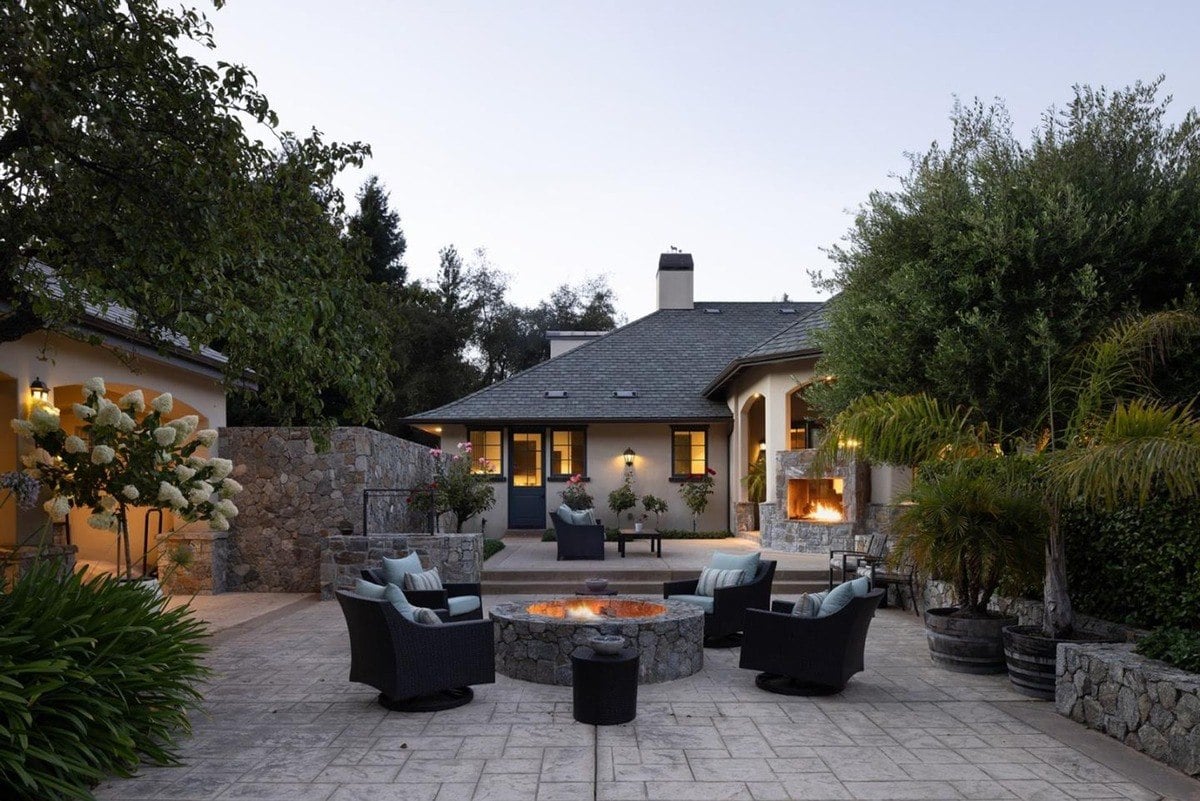 Circular outdoor seating area surrounds a stone firepit amidst a lush patio space.