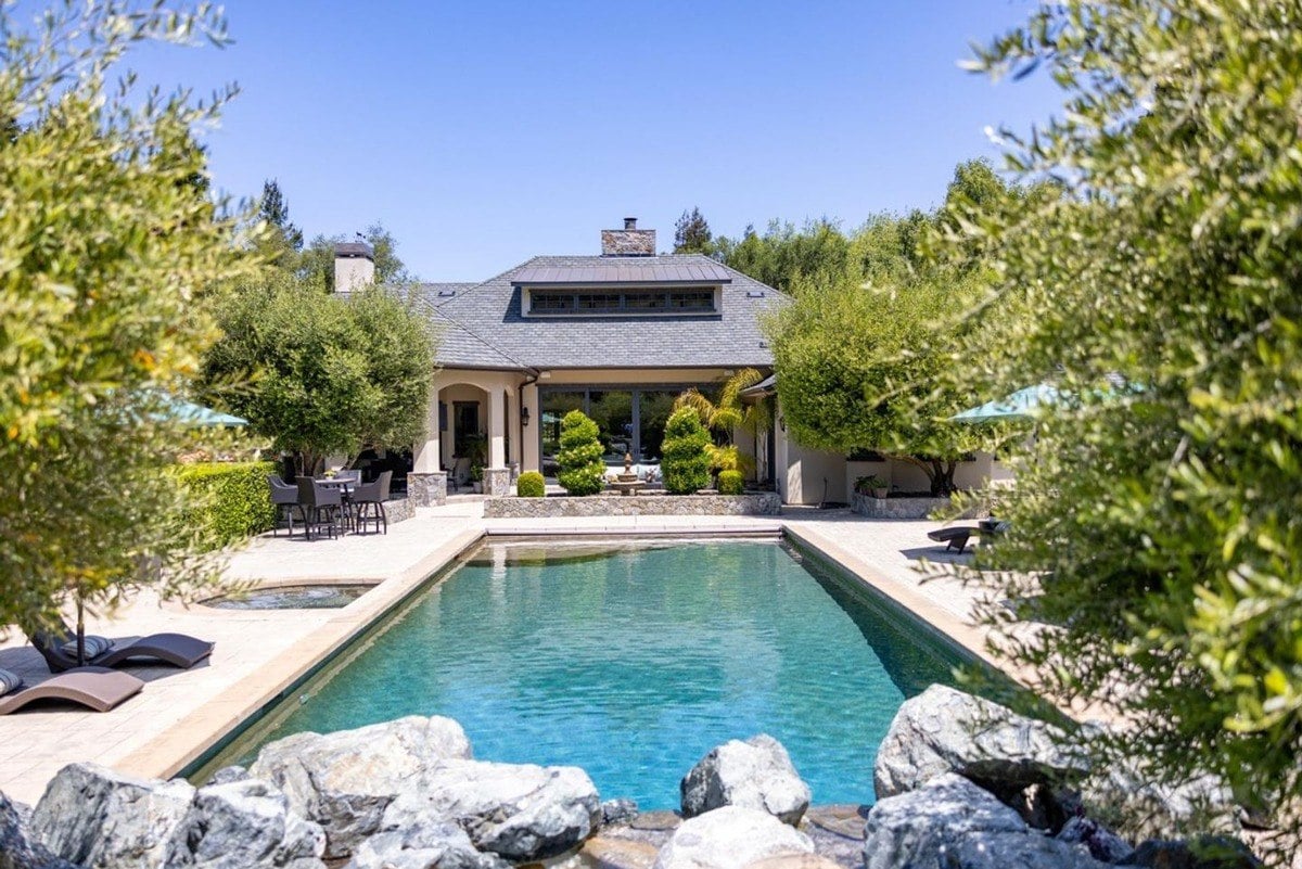 Large pool surrounded by patio seating and lush greenery with a view of the house in the background.