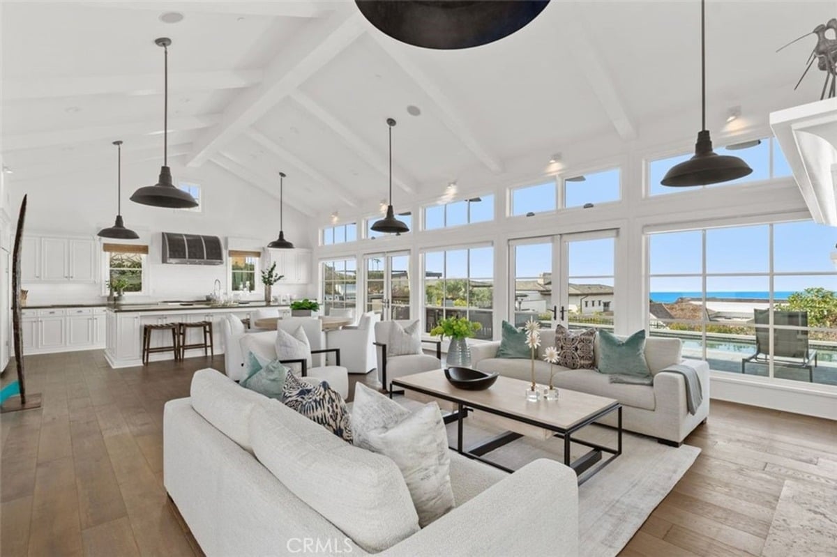 Open-concept living room featuring a high ceiling, multiple black pendant lights, white furniture, and large windows with ocean views.