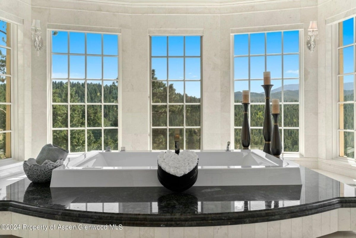 A sleek marble base elevates the freestanding tub, surrounded by tall windows that showcase panoramic forest views.