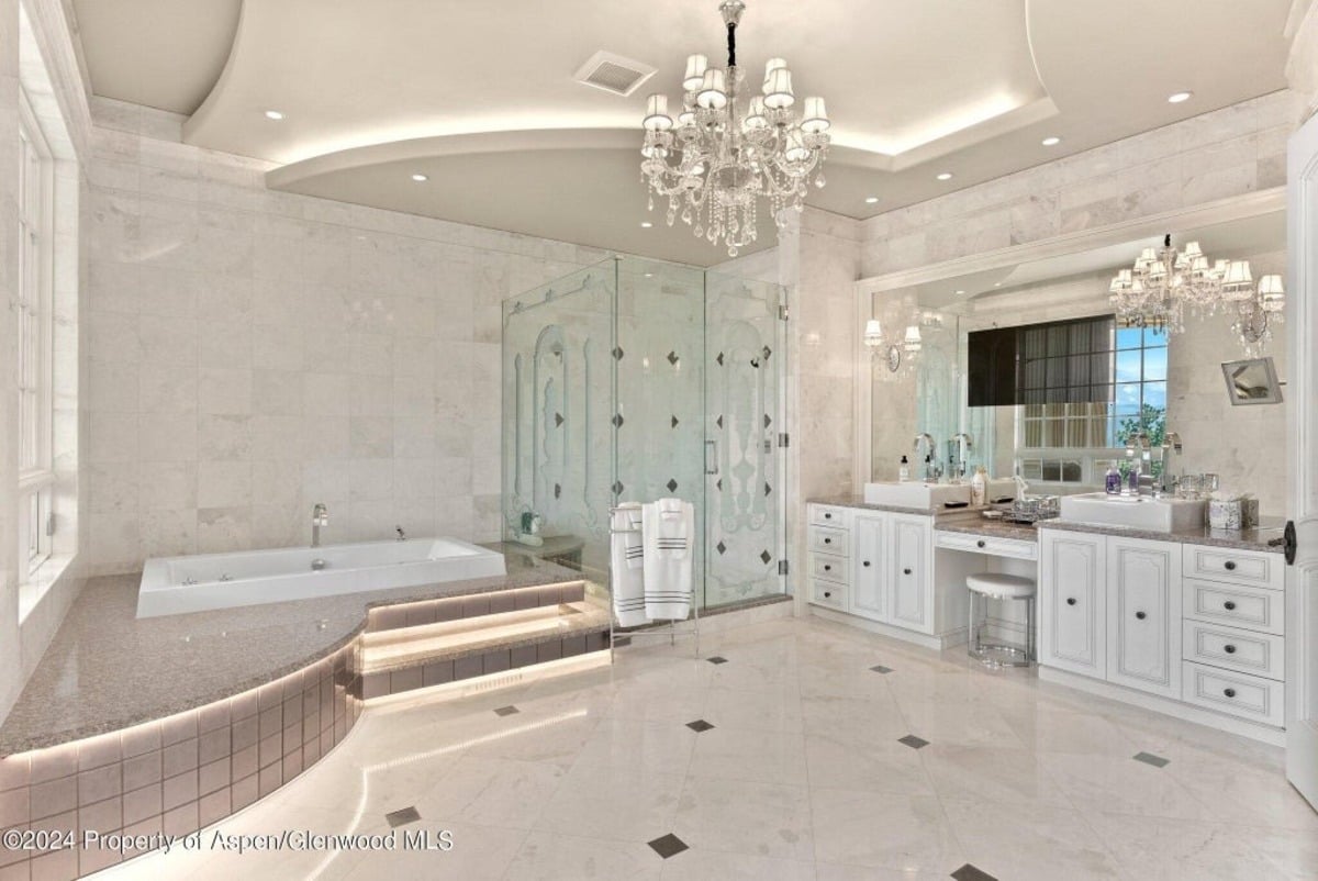 Polished marble walls and floors create an elegant backdrop for the soaking tub set on a raised platform with accent lighting.