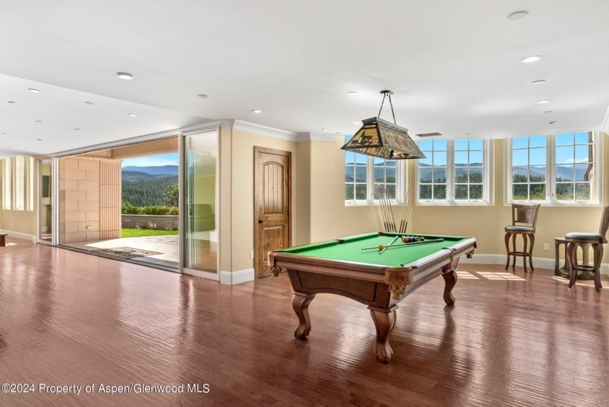 Classic pool table sits beneath a stained-glass billiard light, surrounded by large windows showcasing scenic views.