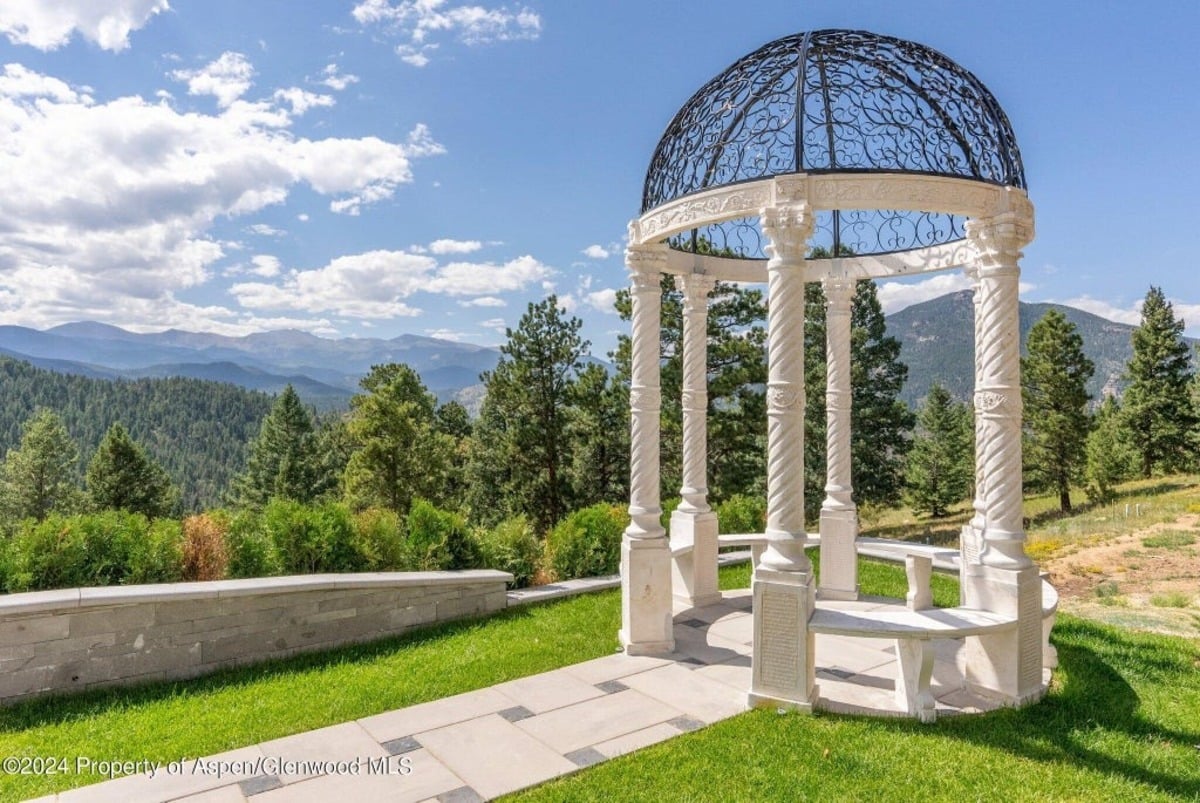 A white stone gazebo with spiral columns and a domed wrought-iron roof sits on a manicured lawn.