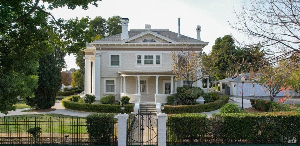 Historic white two-story home features grand columns, a circular porch, and well-manicured hedges behind an iron gate.