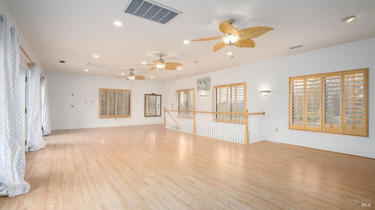 Large recreation room displays hardwood flooring, plantation shutters, and ceiling fans with leaf-shaped blades.