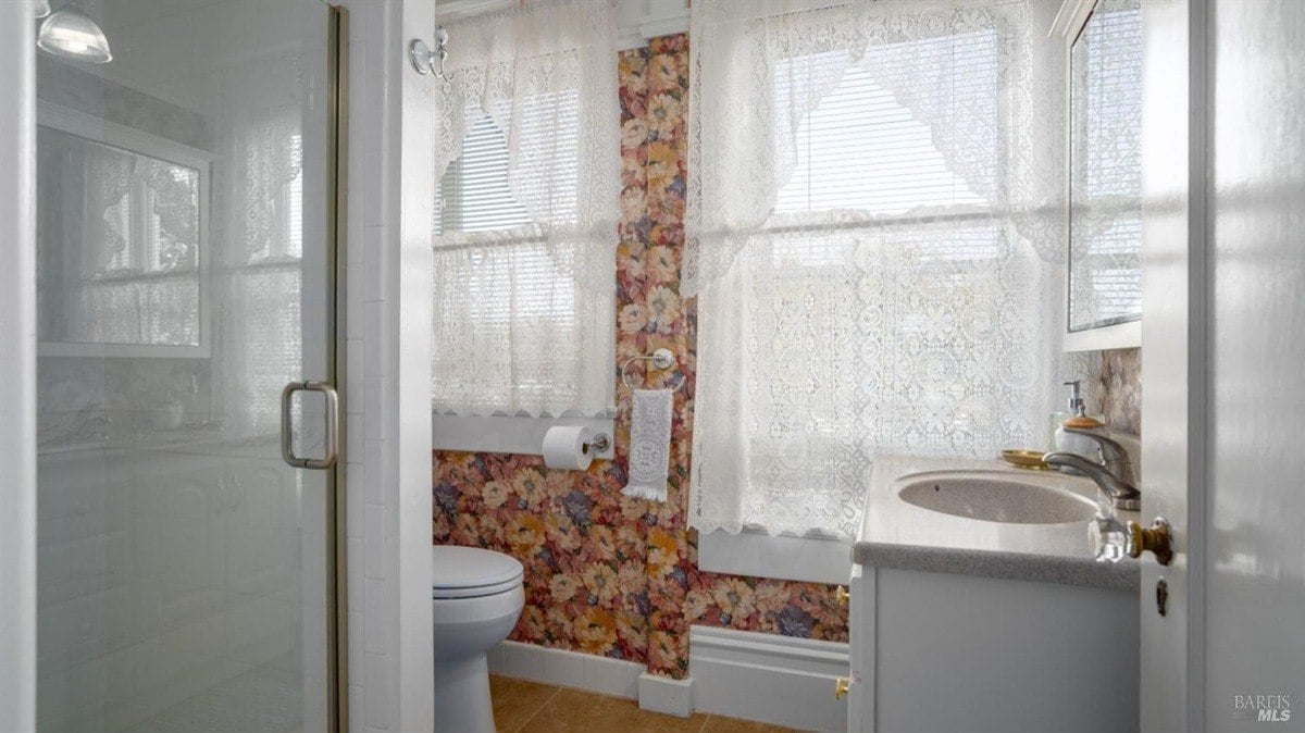 Bright bathroom includes a pedestal sink, floral wallpaper, and sheer lace curtains over the windows.