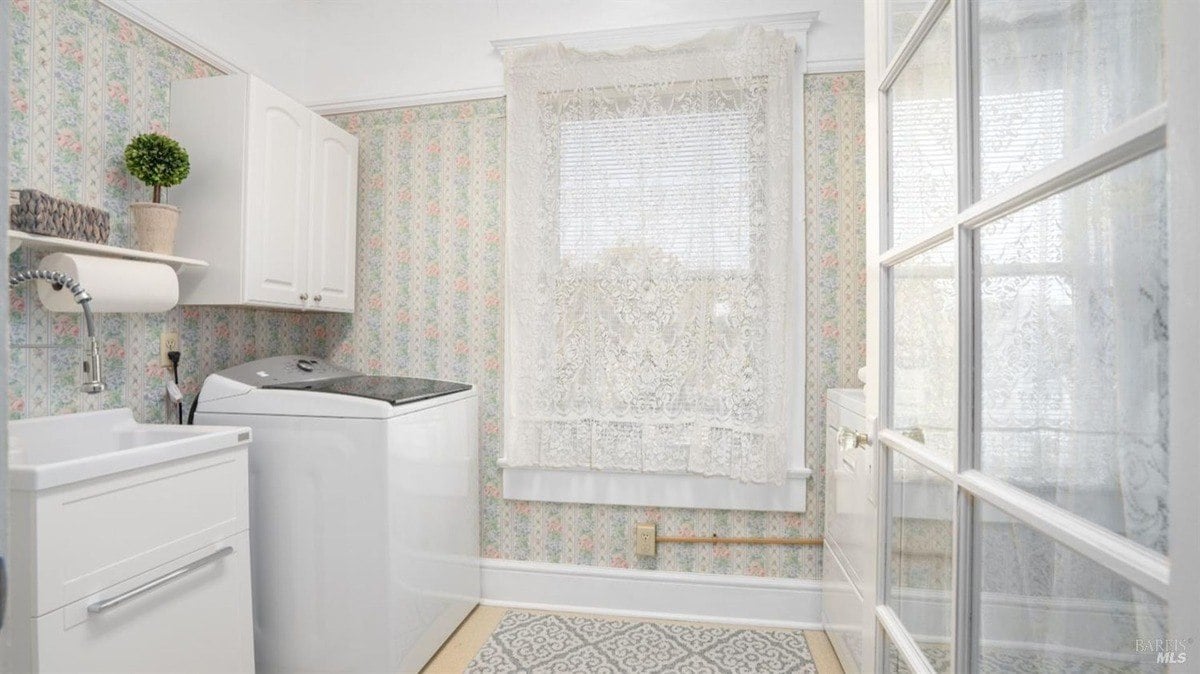 Laundry room contains floral wallpaper, white cabinetry, a washer, and a lace-draped window.