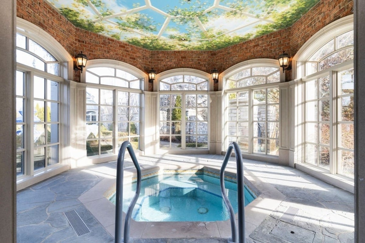 Spa-like space with a hot tub is surrounded by large windows and a ceiling mural of greenery and sky.