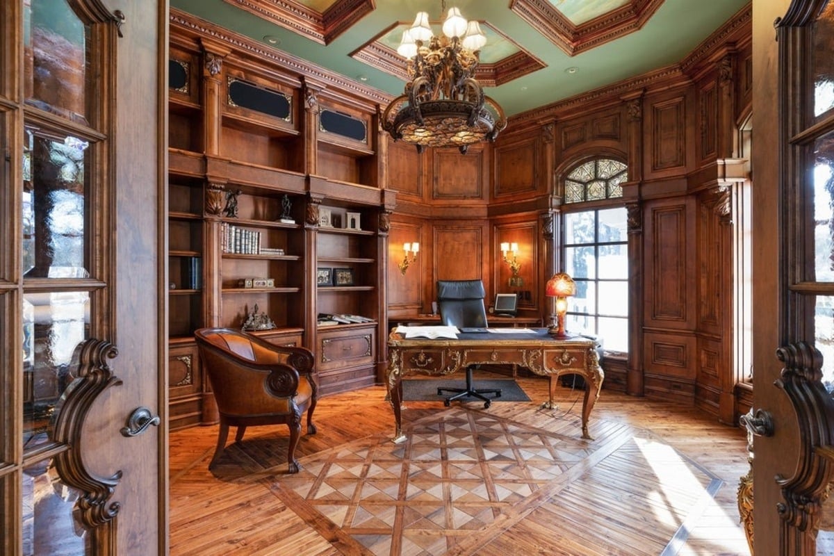 Office with floor-to-ceiling wood paneling, built-in bookshelves, and a central antique desk under an elaborate chandelier.
