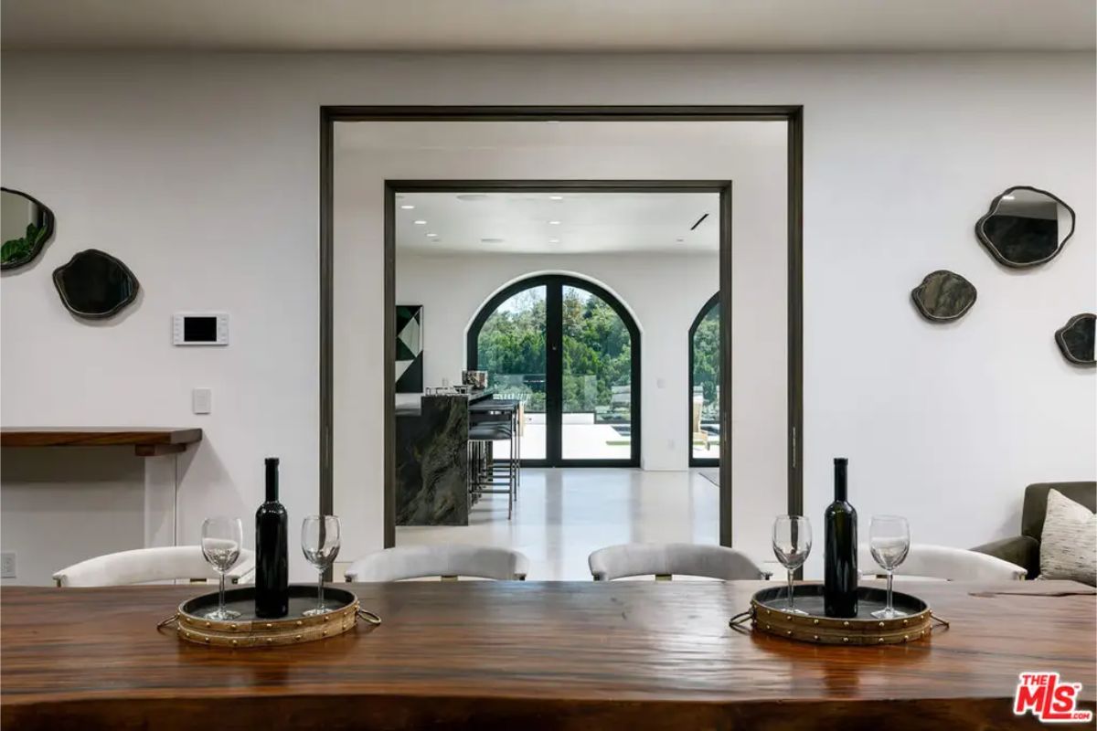Dining room view features a rustic wooden table with wine bottles and glasses, framed by an arched doorway revealing a bright room beyond. 
