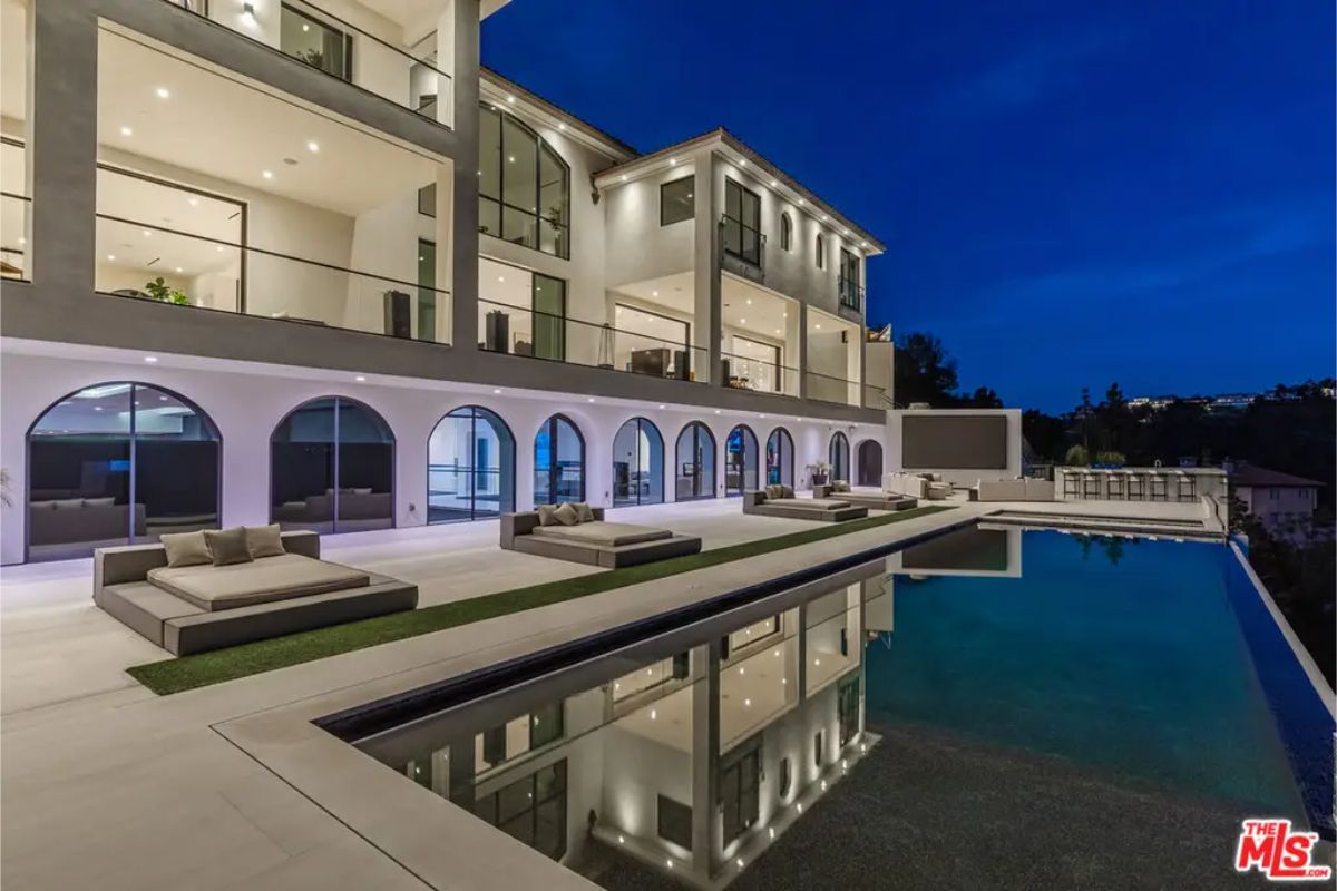 Luxurious poolside area with modern loungers, reflecting the multi-story glass facade of the mansion at dusk. 