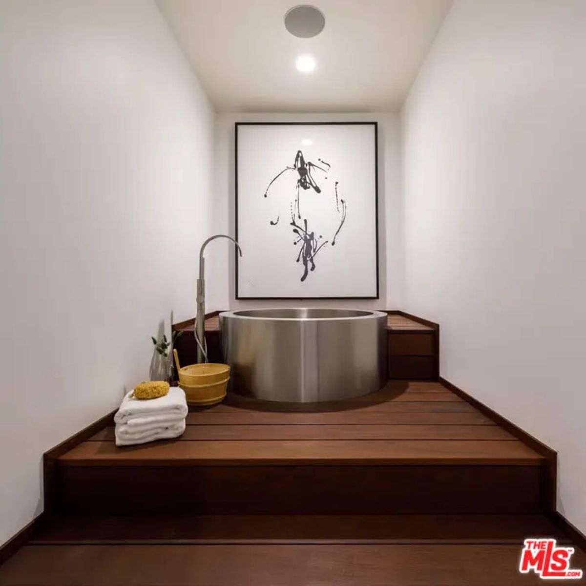 Minimalist bathing area with a stainless steel soaking tub on a wooden platform, complemented by modern artwork. 
