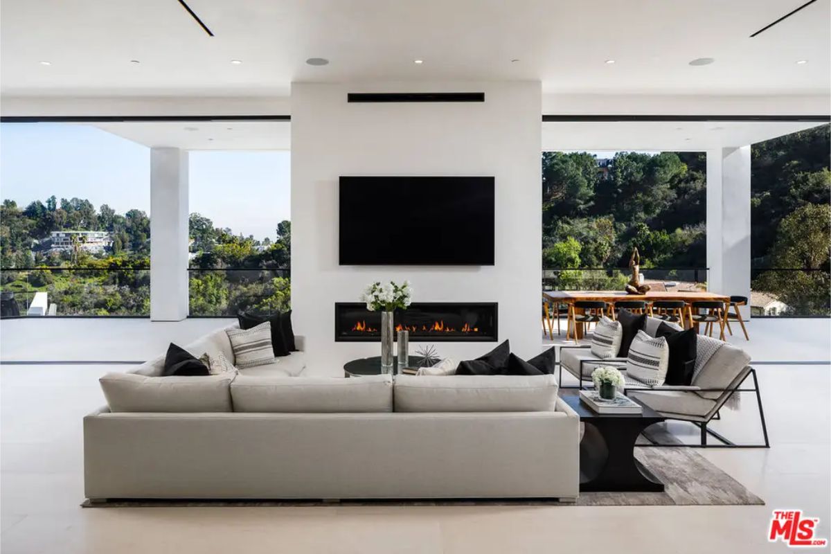 Modern living room showcases a sleek fireplace beneath a wall-mounted TV, complemented by a spacious sectional sofa and floor-to-ceiling windows with scenic views. 