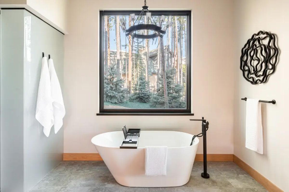 Freestanding bathtub with a black fixture, large picture window, and modern chandelier. 
