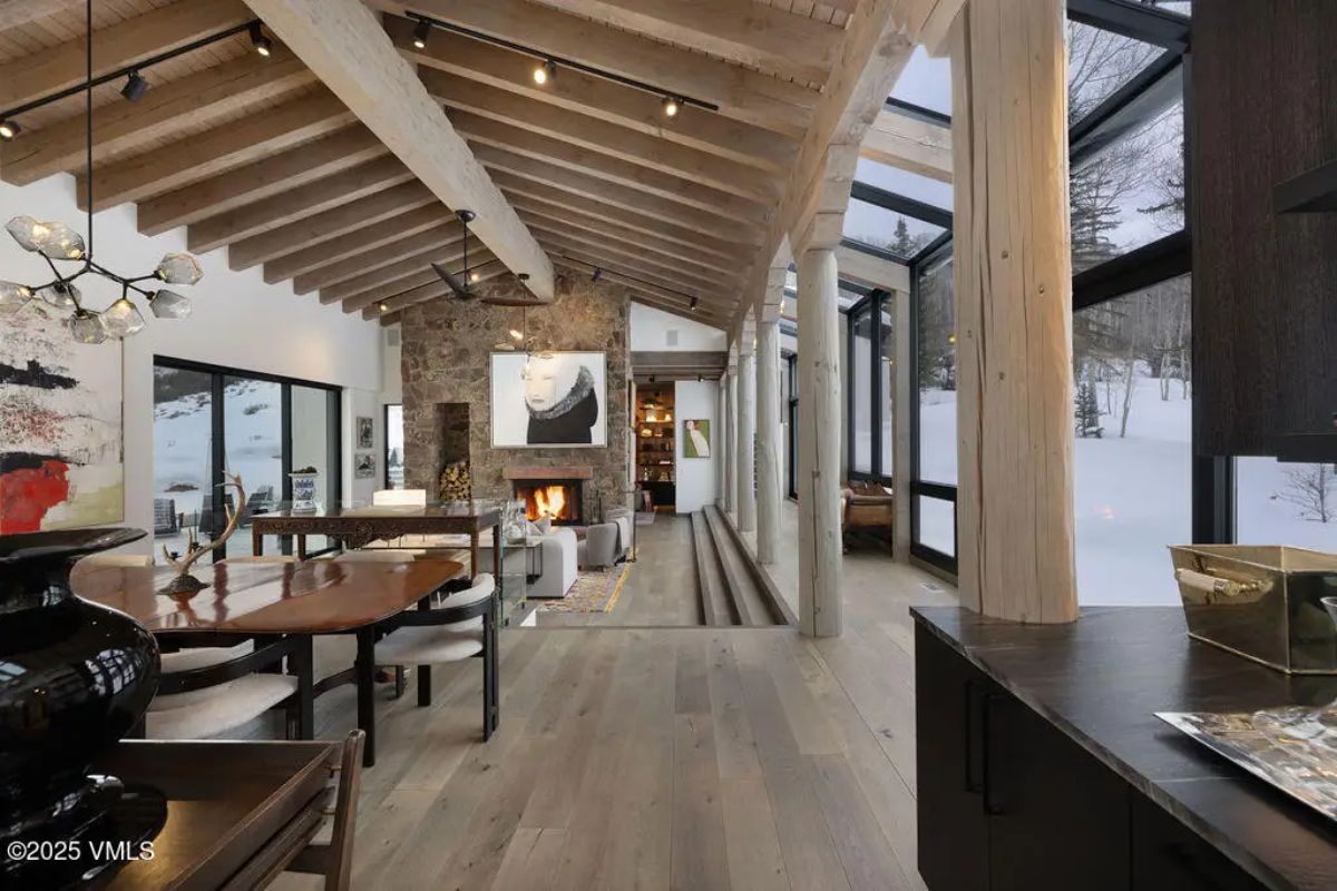 Open-concept living and dining area with exposed wood beams, floor-to-ceiling windows, and a stone fireplace as the focal point.