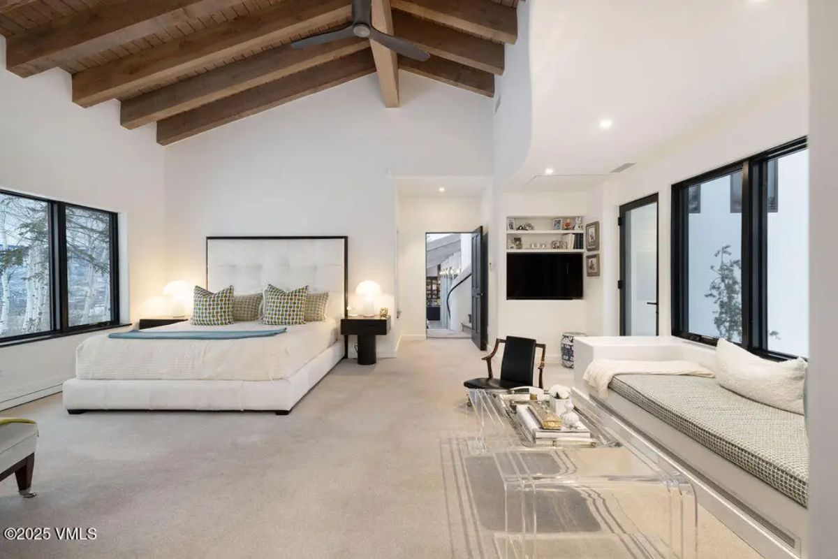 Exposed wooden beams, large windows offering natural light, and neutral-toned carpeting.