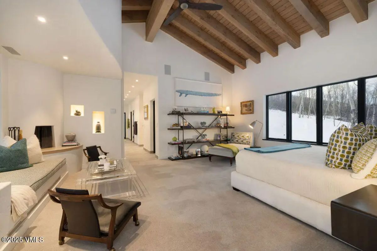 Bedroom with exposed wooden beams, large windows overlooking a snowy landscape, and neutral carpeting.