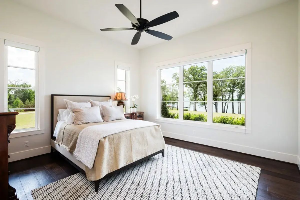 Bedroom with white walls, dark hardwood flooring, and large windows overlooking a lake and trees.
