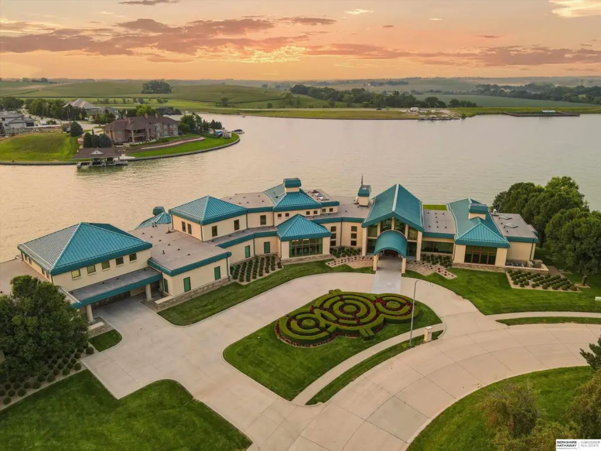 Front-facing aerial perspective of the grand estate, showing its stunning lakefront location, manicured lawn, and curved driveway with an intricate garden design. 
