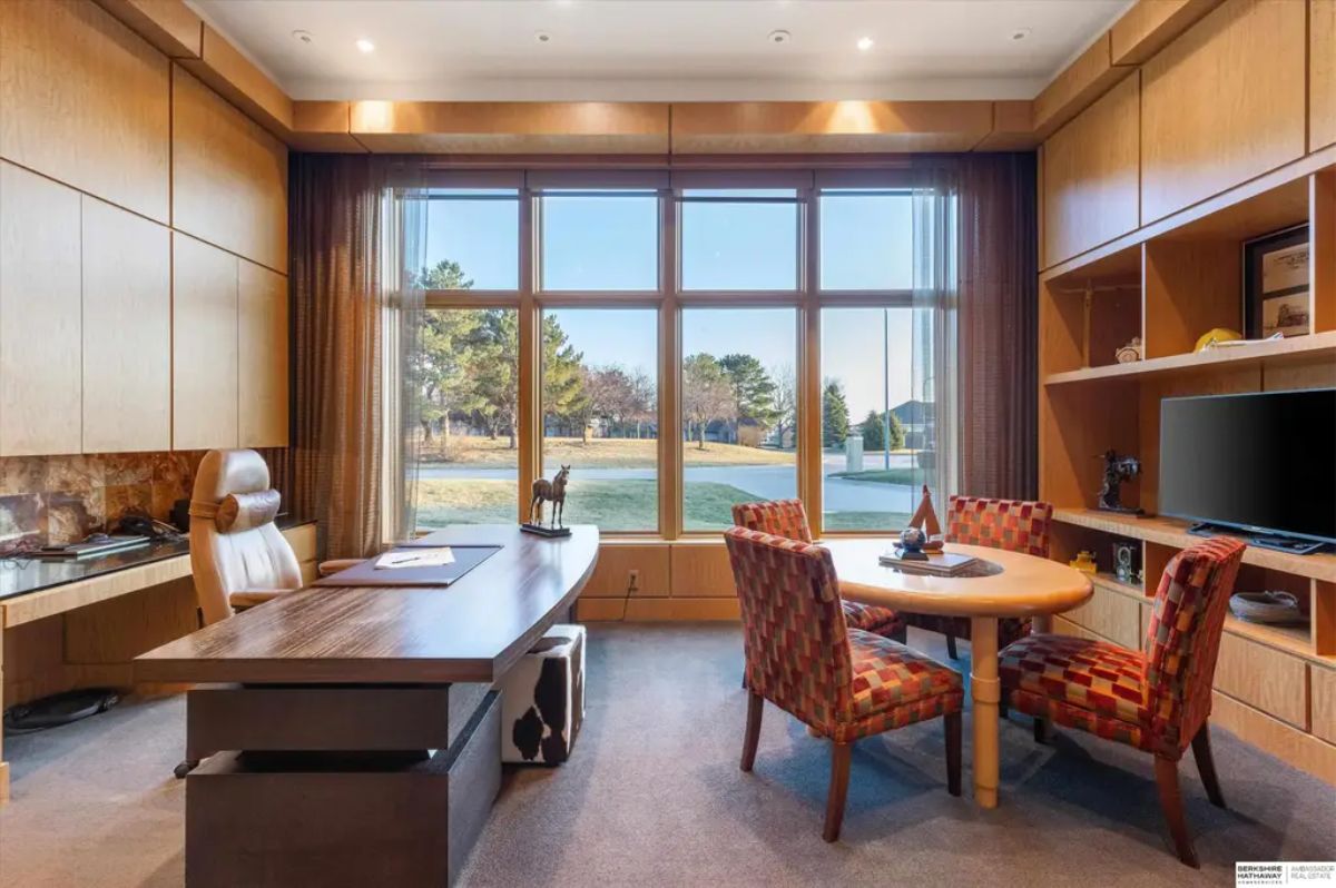 Luxurious home office featuring wood paneling, a sleek desk, and a cozy meeting area with a large window letting in natural light. 