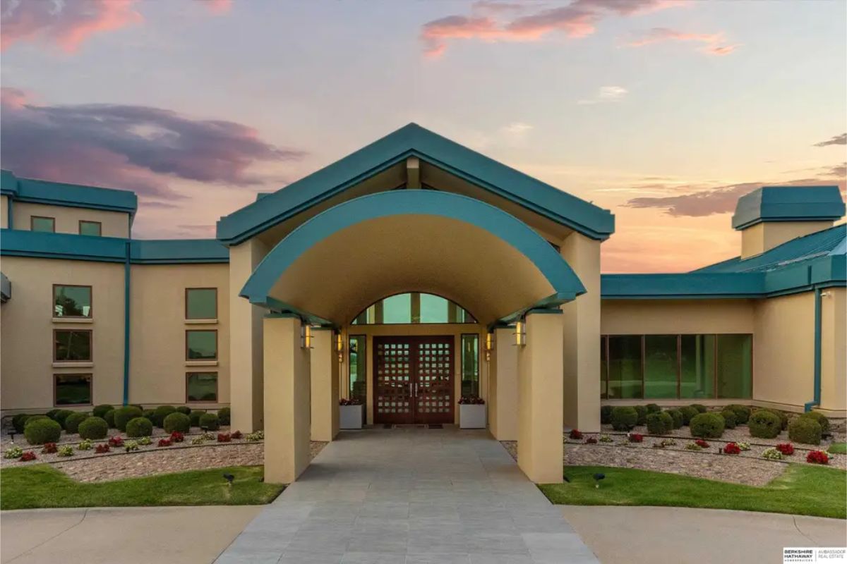 Grand entrance to the estate with an arched entryway, elegant lighting, and meticulously maintained landscaping set against a sunset backdrop. 