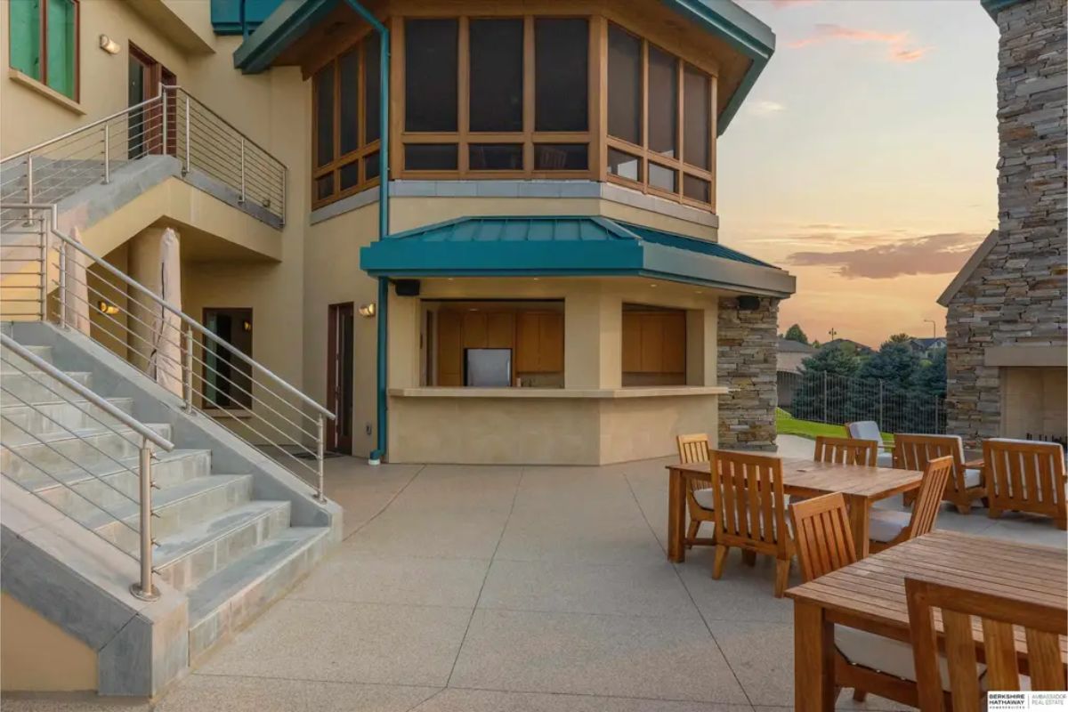 Outdoor bar and dining space showcases wooden patio furniture, a stone fireplace, and a well-designed covered bar area near a grand staircase. 