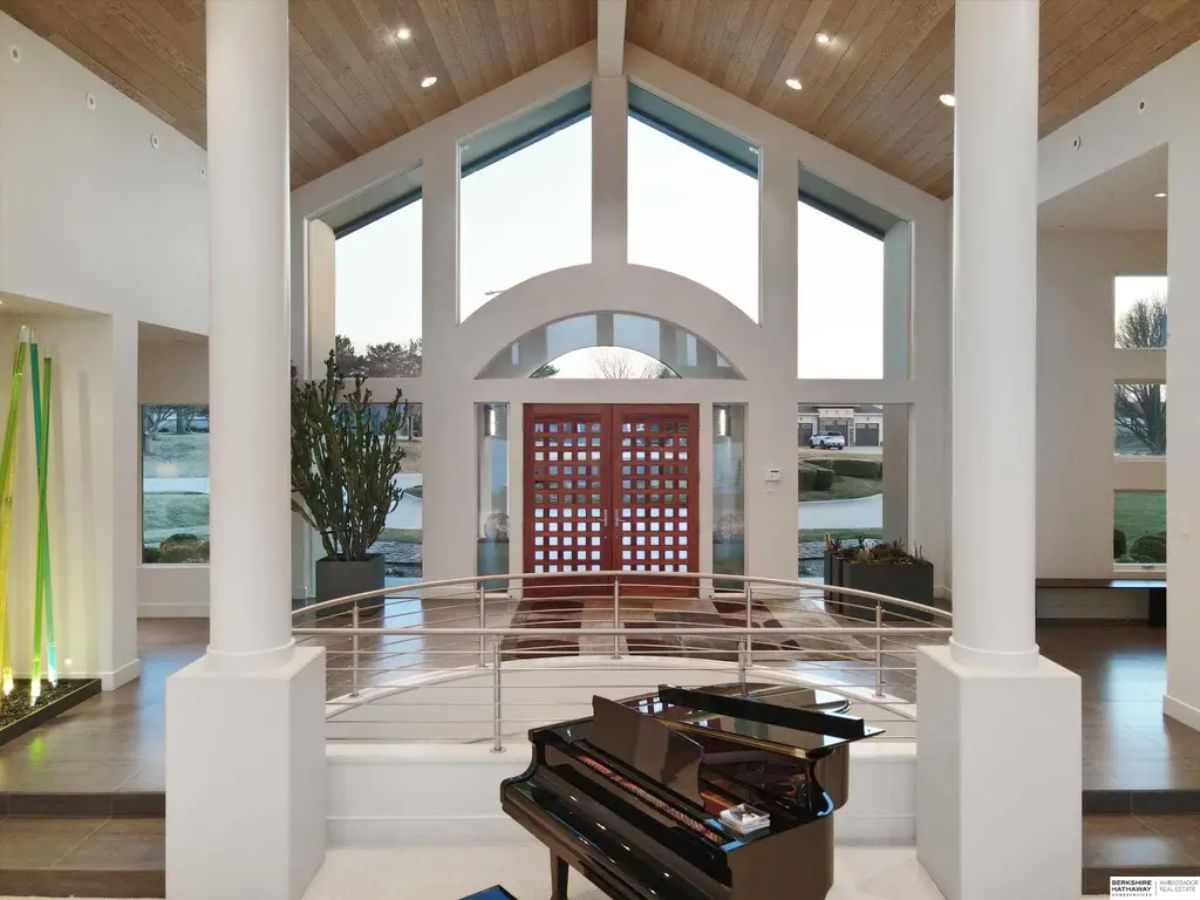 Interior foyer featuring high ceilings, a curved staircase, and a sleek grand piano, with large windows letting in abundant natural light. 