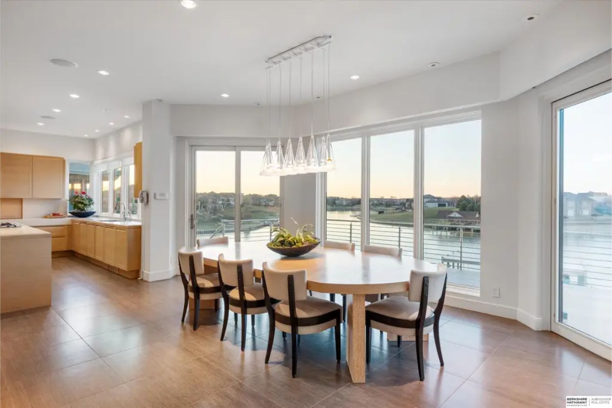 Dining area with floor-to-ceiling windows, a round wooden table, and pendant lighting, overlooking a scenic lake view at sunset. 